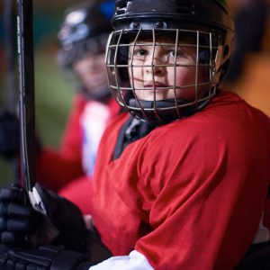 Picture of child sitting on bench with red ice hockey jersey holding hockey stick and black helment